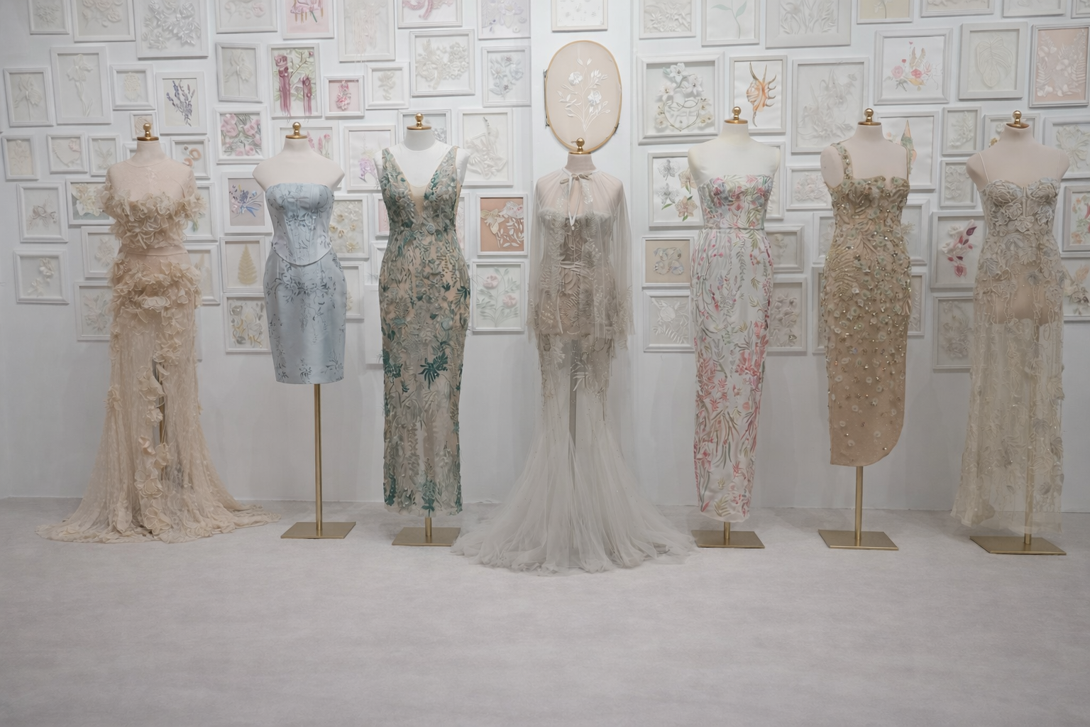 Seven made-to-order gowns displayed on mannequins in front of a wall of framed embroidery sketches, Hermione de Paula Made To Order